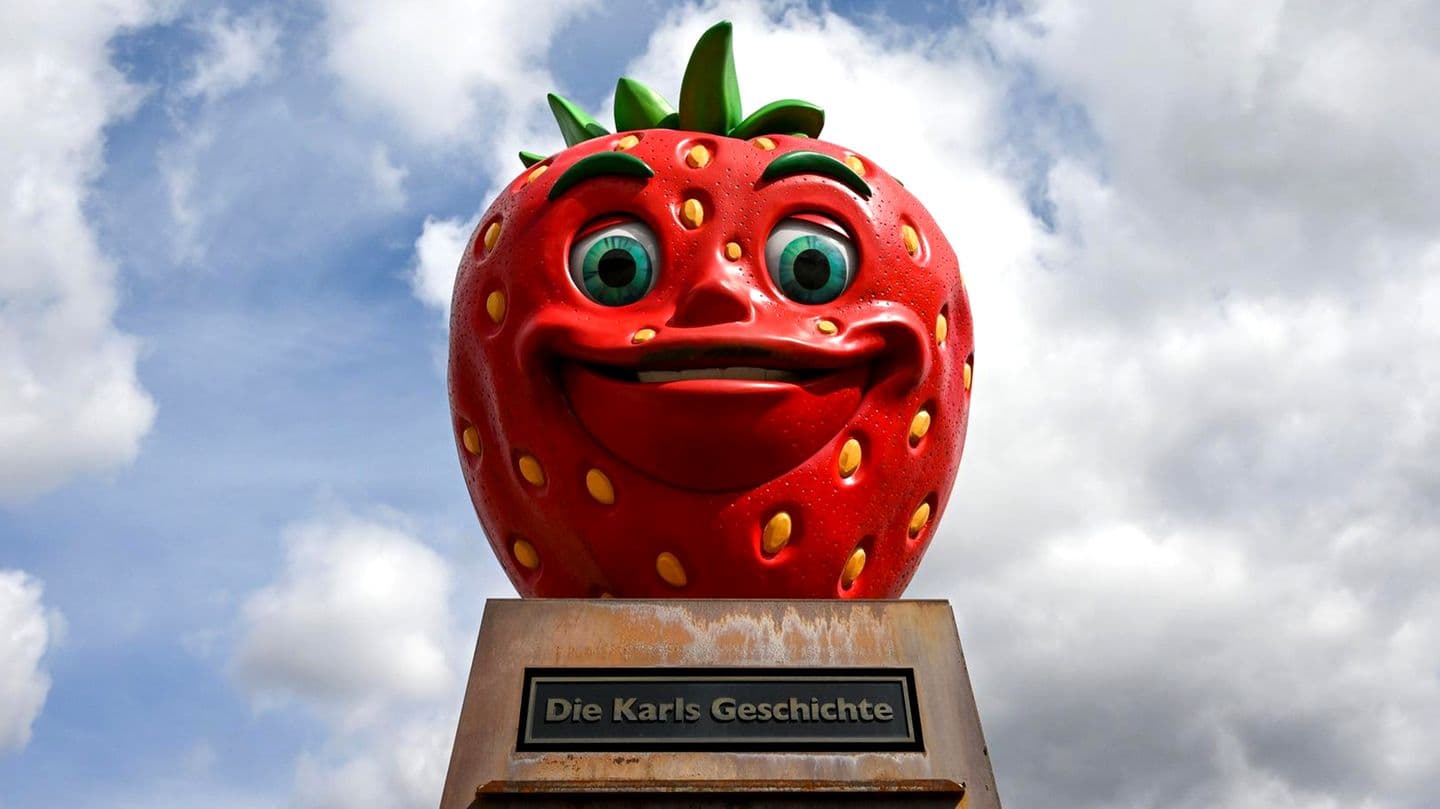 Giant smiling strawberry statue with green leaves on top, set against a cloudy sky. Plaque reads "Die Karls Geschichte."