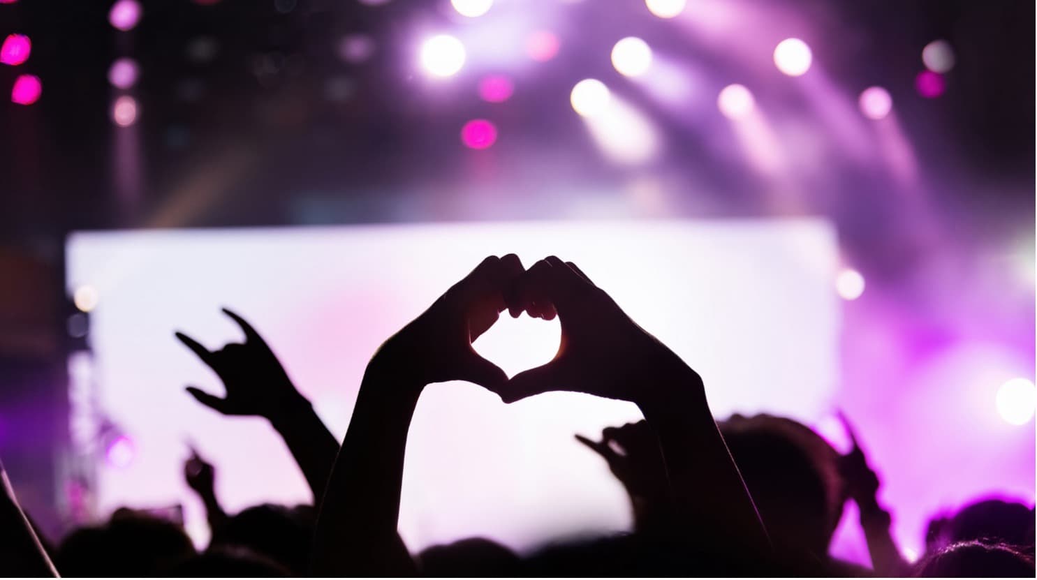 Silhouetted hands forming a heart at a pink-purple lit concert stage, crowd and rock-hand gestures in the foreground.