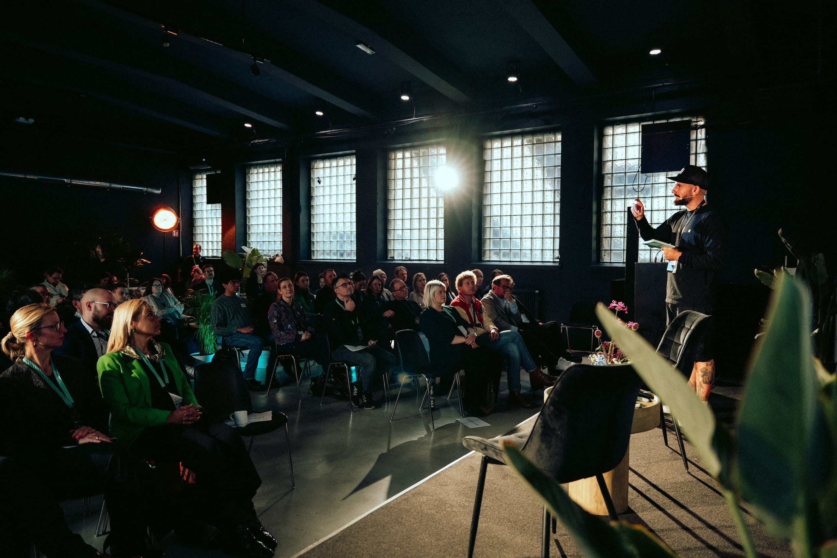 A speaker addresses a seated audience in a dimly lit room with large windows and spotlights. The audience listens attentively.