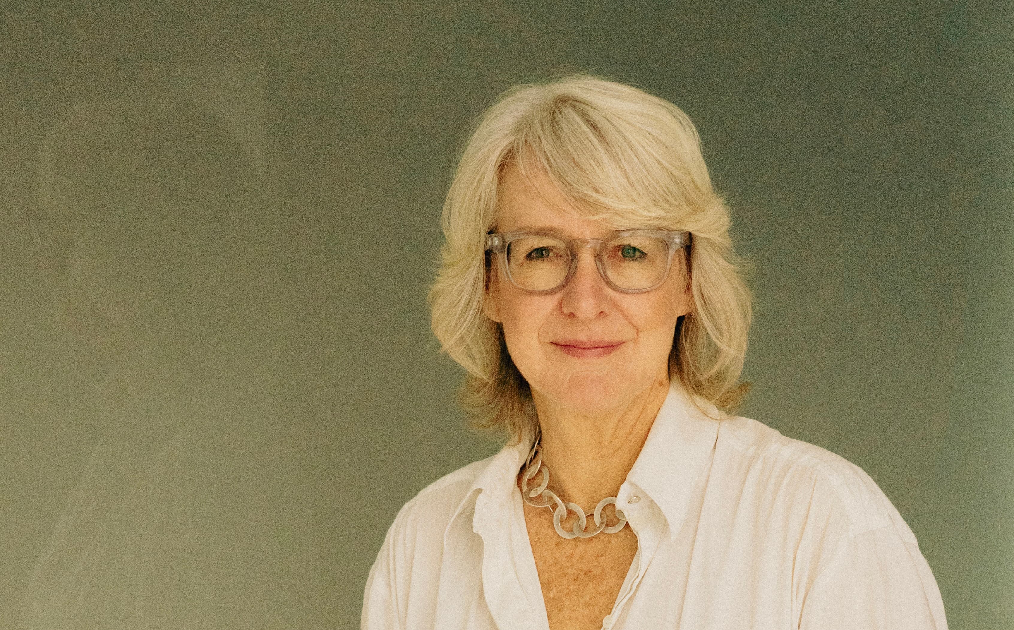 Woman with short gray hair and glasses, wearing a white shirt and silver necklace, smiling against a plain background.