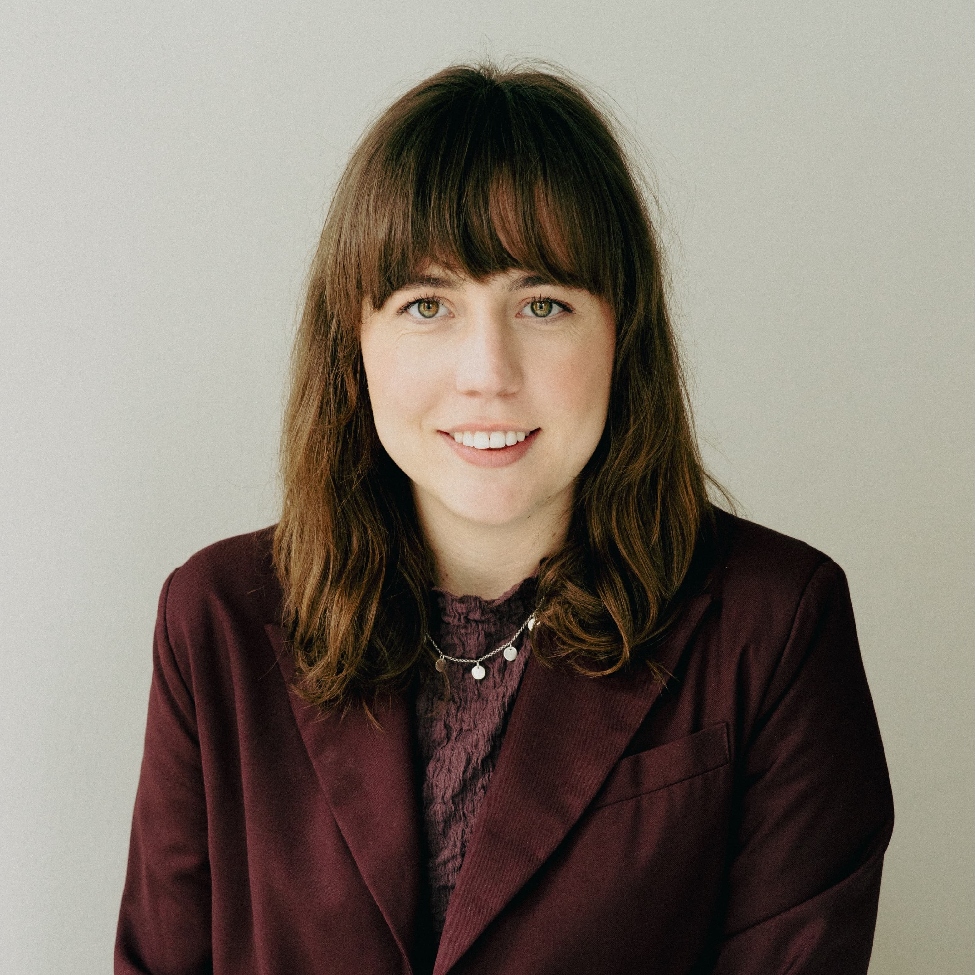 Woman with shoulder-length brown hair in a maroon blazer, smiling at the camera against a plain background.