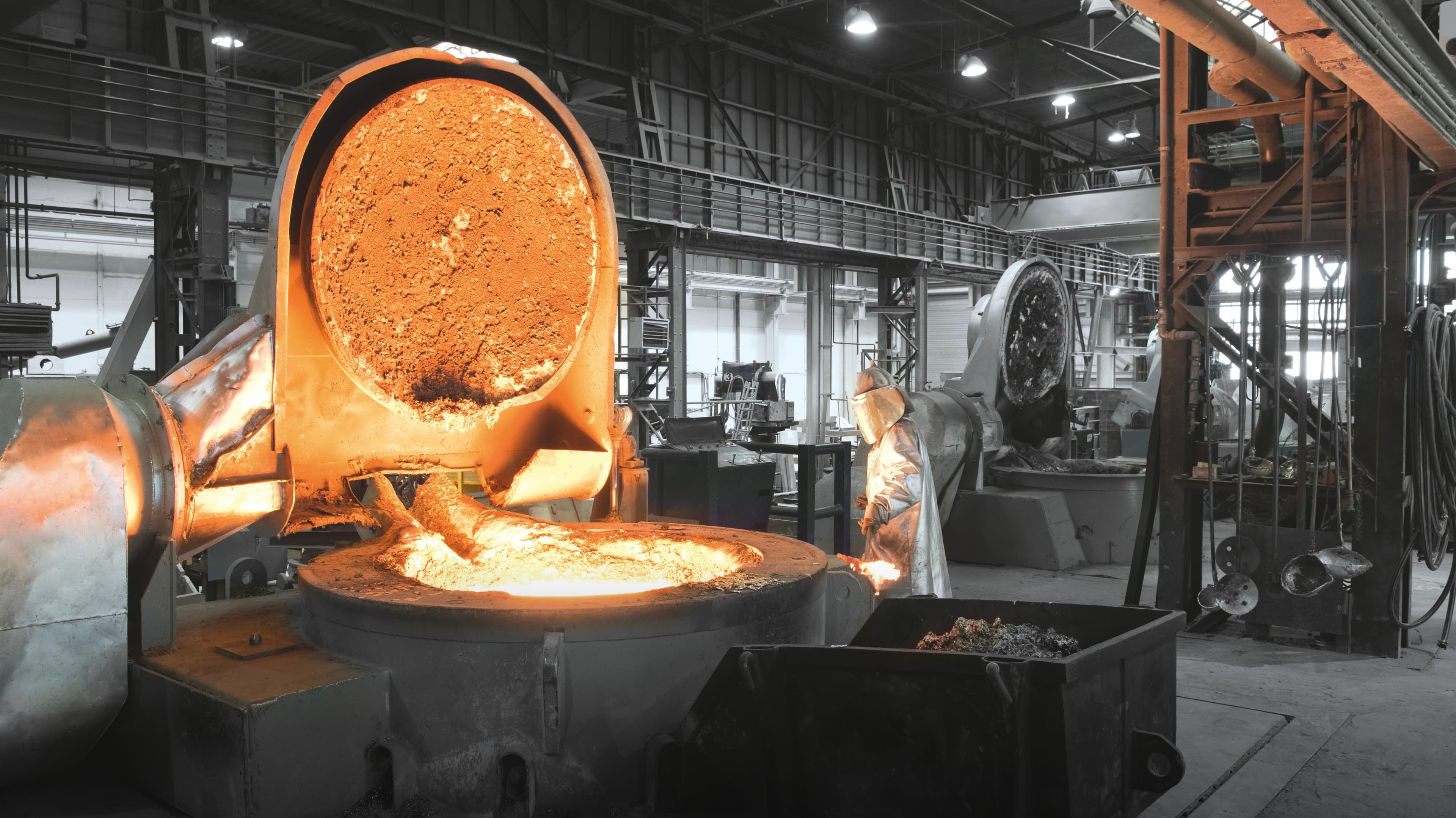Worker in protective gear observes molten metal in a large industrial furnace within a spacious factory setting.