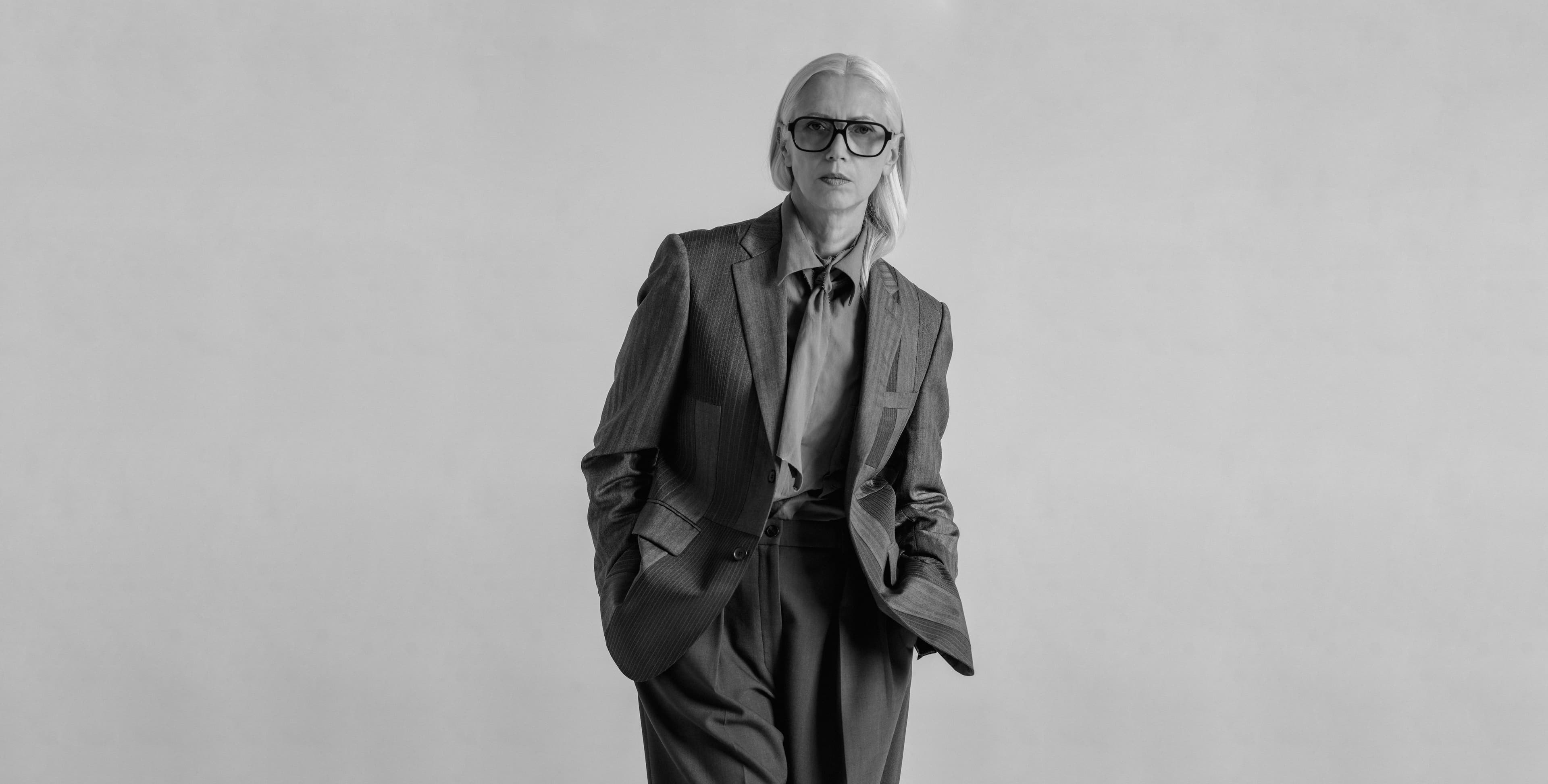 Black and white photo of a person wearing glasses and a suit, hands in pockets, looking confidently at the camera.